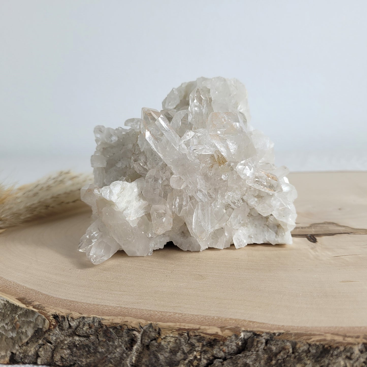 Bergkristall Naturstufe mit klaren Kristallen, platziert auf einer Holzscheibe, neutraler Hintergrund.