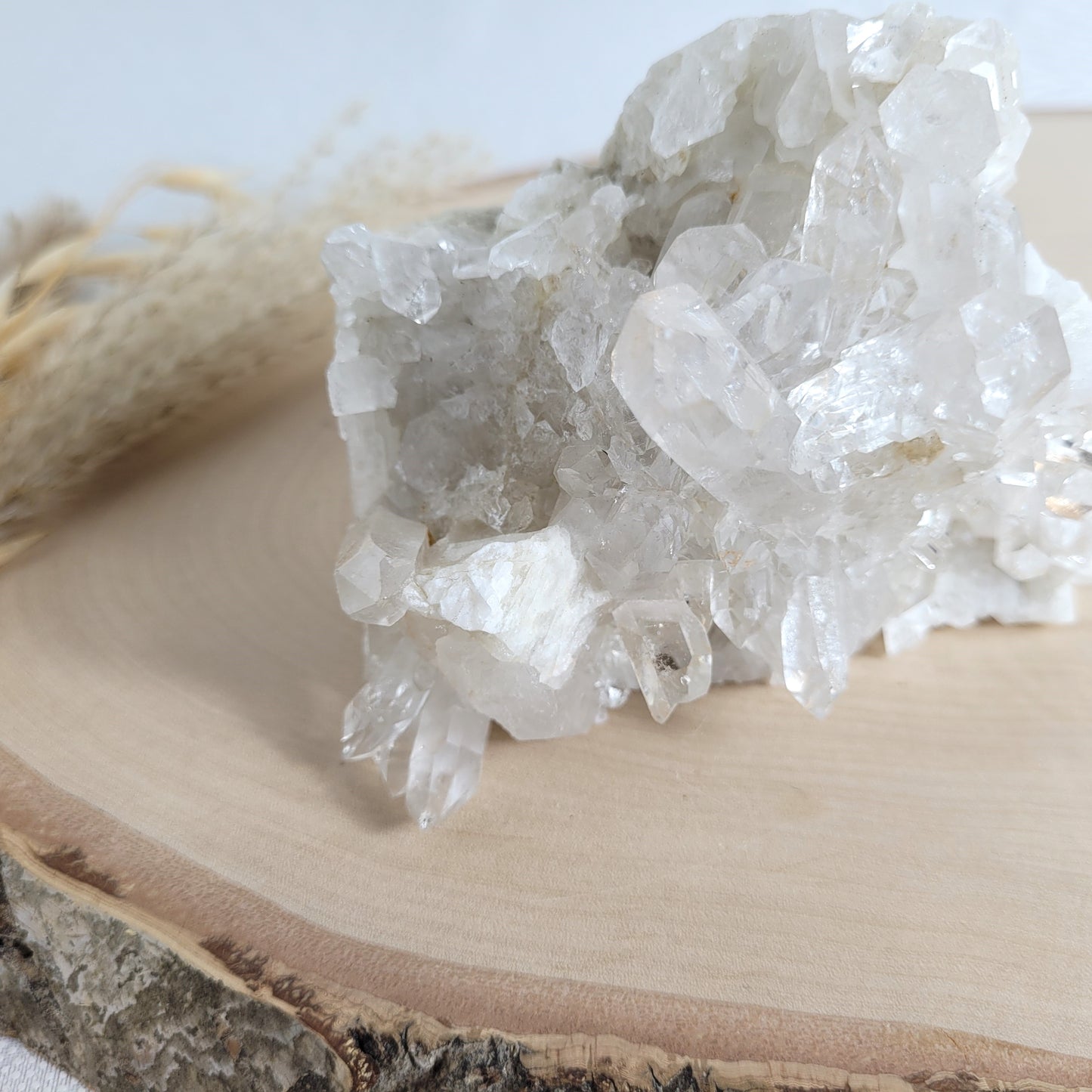 Bergkristall Naturstufe mit klaren Kristallen, platziert auf einer Holzscheibe, neutraler Hintergrund.