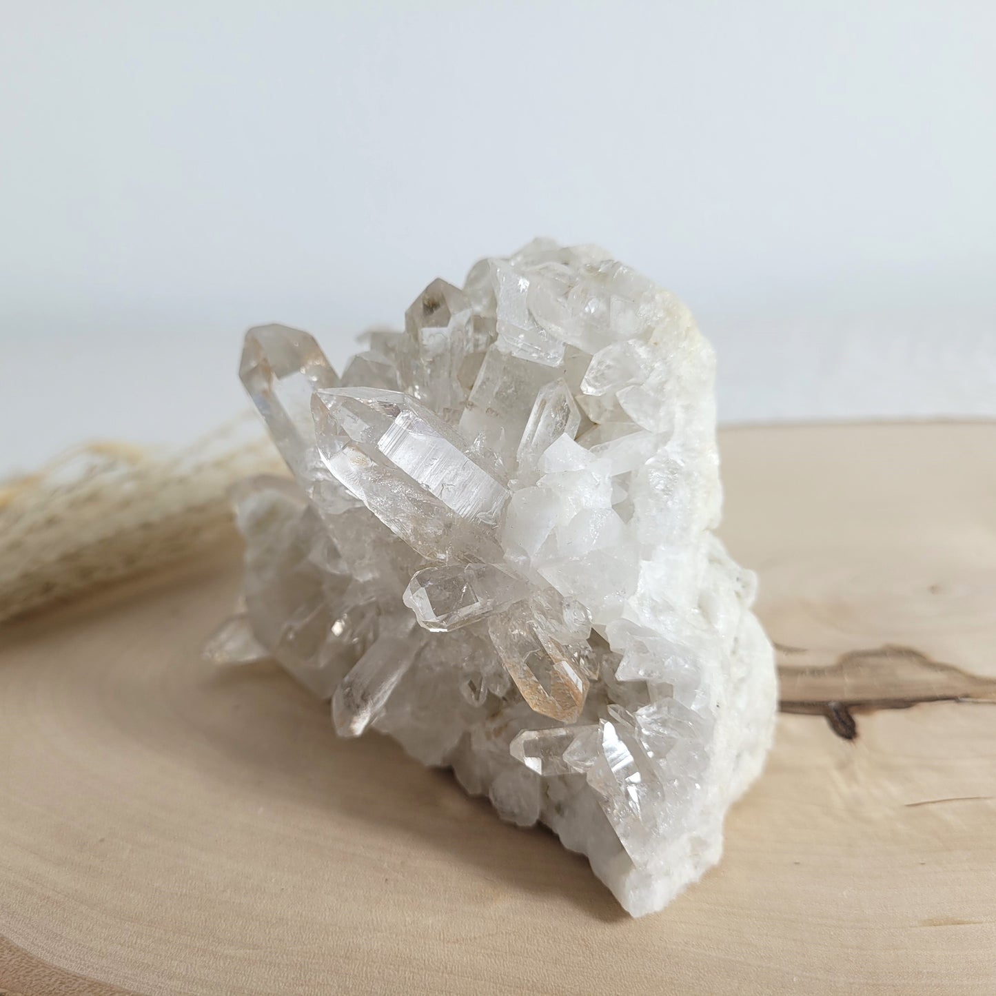 Bergkristall Naturstufe mit klaren Kristallen, platziert auf einer Holzscheibe, neutraler Hintergrund.