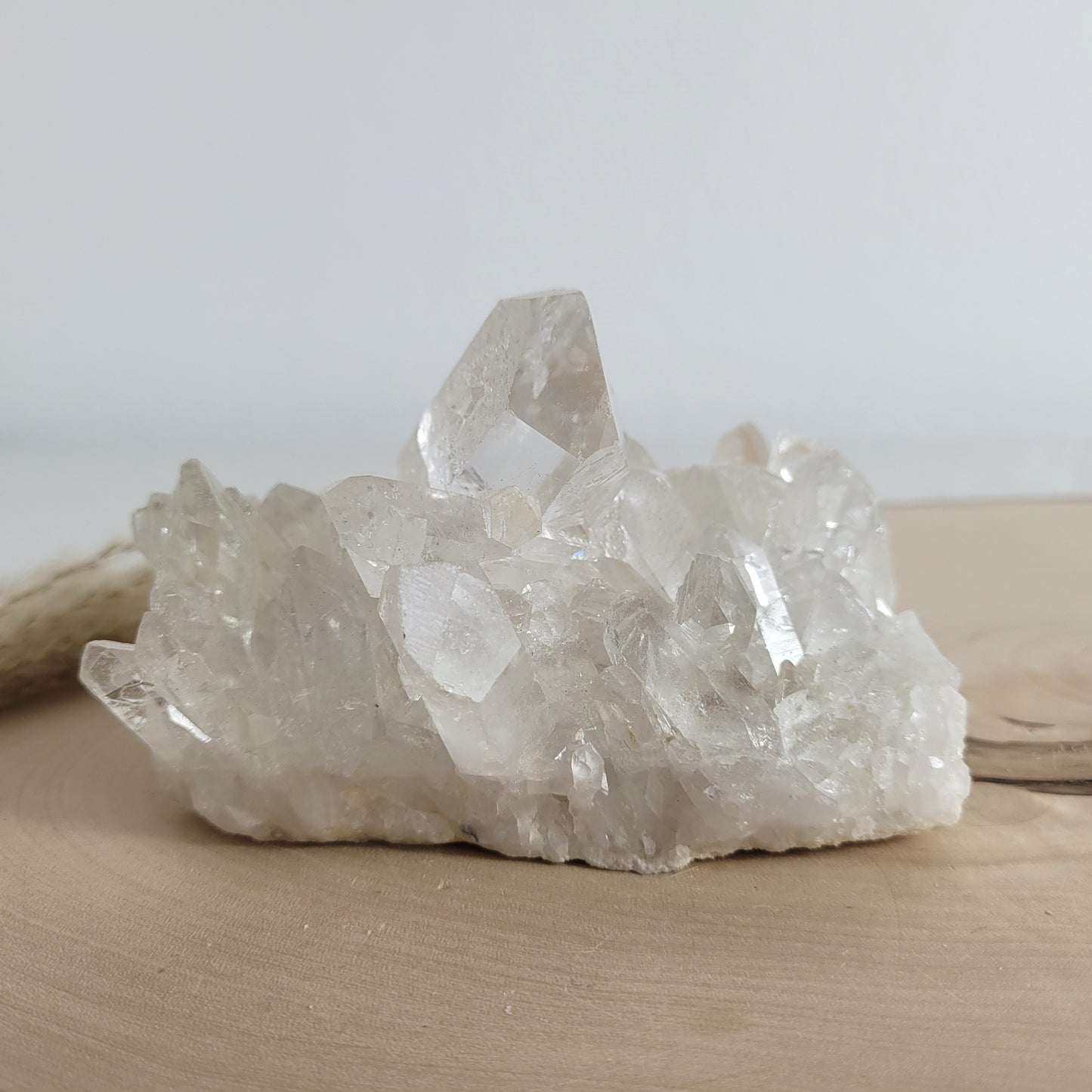Bergkristall Naturstufe mit klaren Kristallen, platziert auf einer Holzscheibe, neutraler Hintergrund.