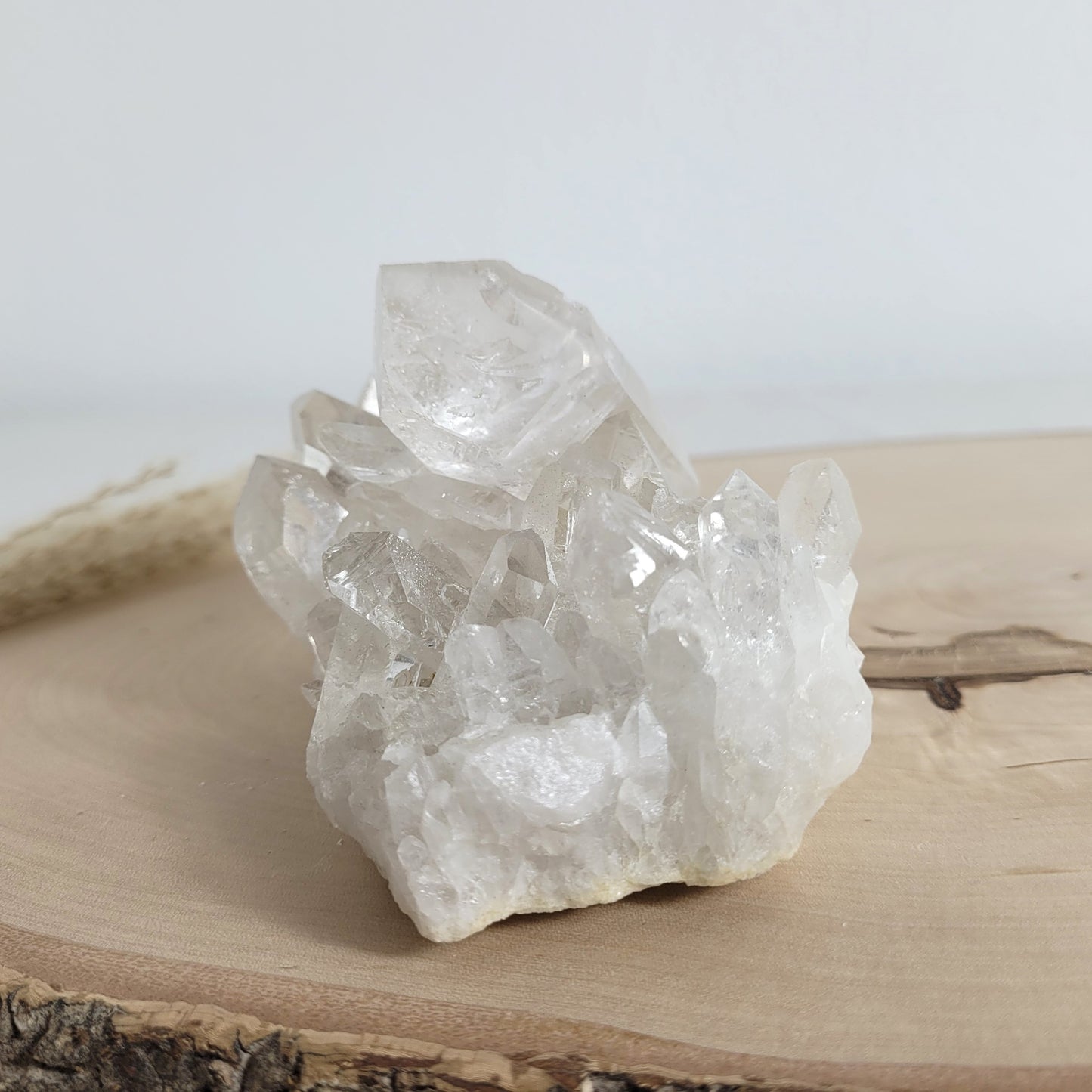 Bergkristall Naturstufe mit klaren Kristallen, platziert auf einer Holzscheibe, neutraler Hintergrund.