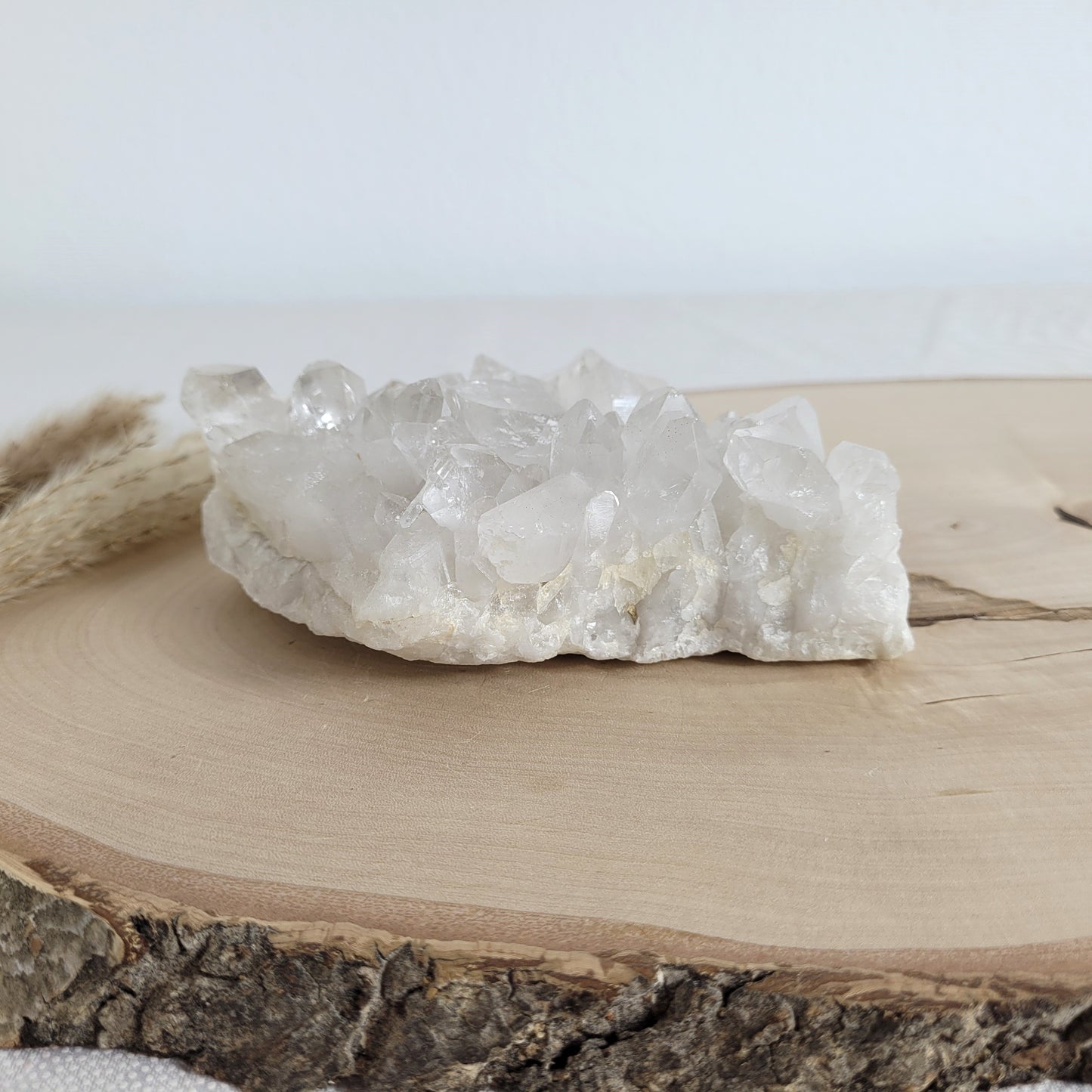 Bergkristall Naturstufe mit klaren Kristallen, platziert auf einer Holzscheibe, neutraler Hintergrund.