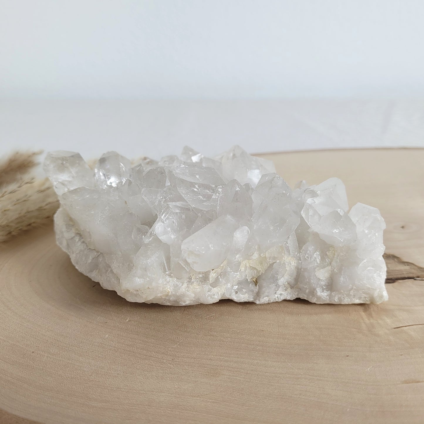 Bergkristall Naturstufe mit klaren Kristallen, platziert auf einer Holzscheibe, neutraler Hintergrund.