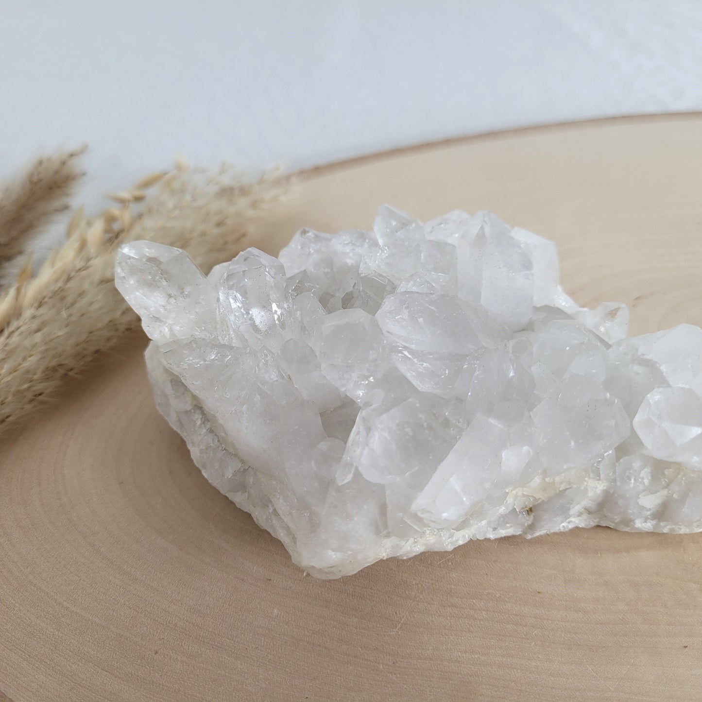 Bergkristall Naturstufe mit klaren Kristallen, platziert auf einer Holzscheibe, neutraler Hintergrund.