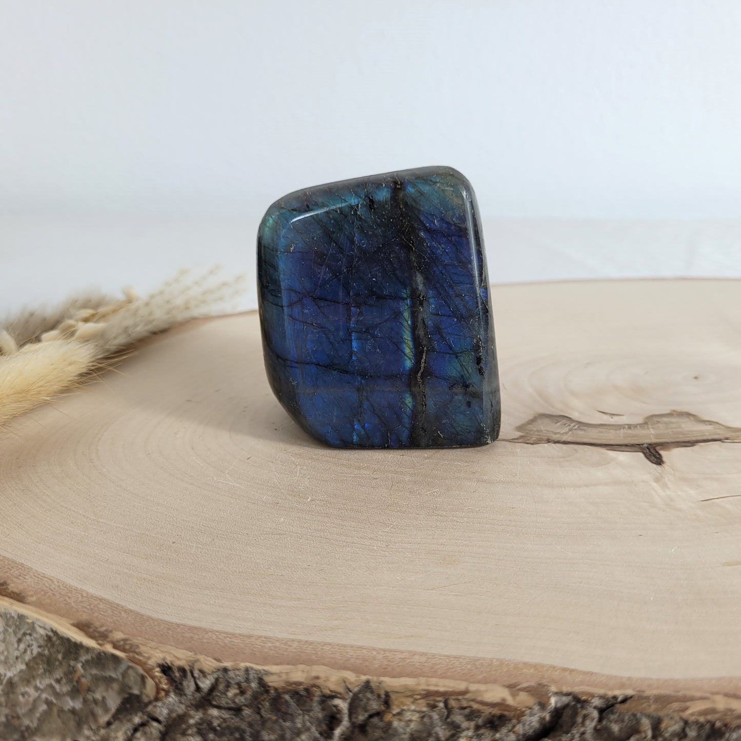 Labradorit Freiform mit natürlichem, intensivem Farbspiel, präsentiert auf einer Holzscheibe. Die typische Lamellenstruktur bricht das Licht auf besondere Weise und macht das Stück zu einem besonderen Blickfang.