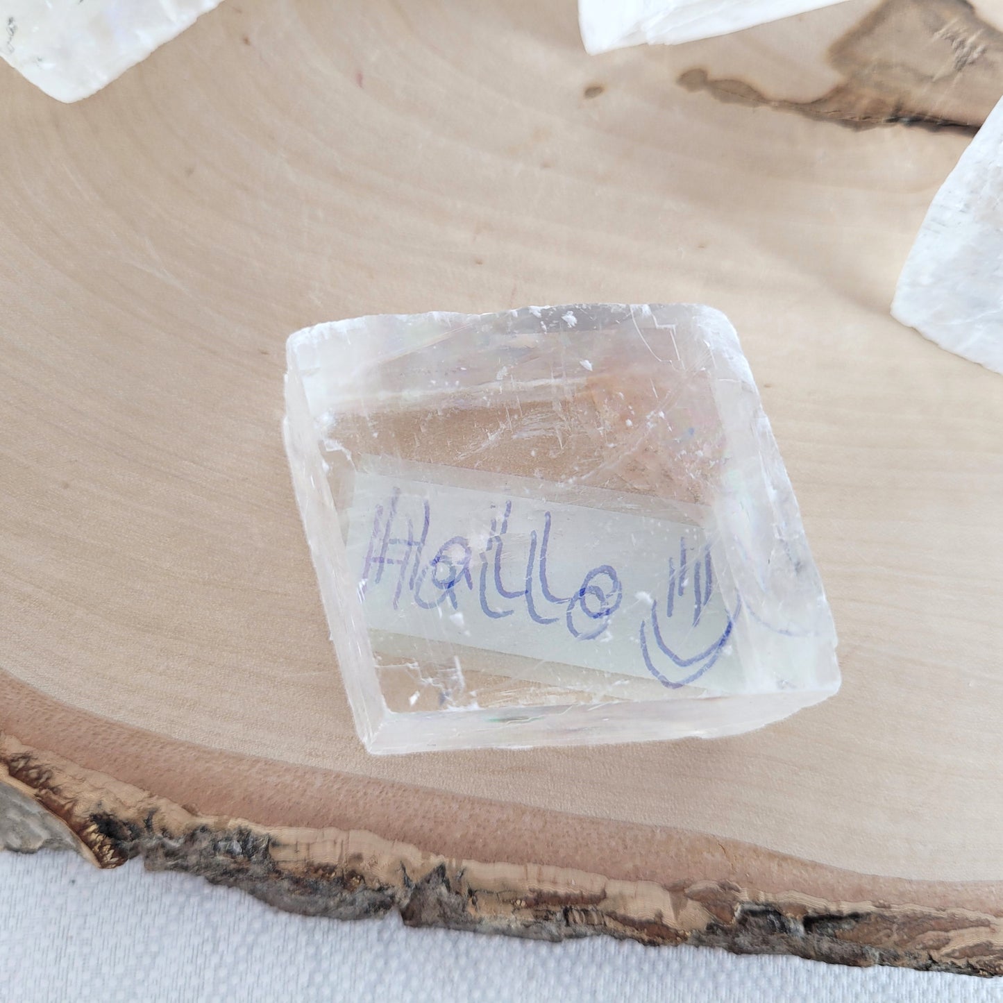 Doppelspat Kristall auf einer runden Holzscheibe präsentiert, naturbelassen mit sichtbarer Maserung – der Kristall zeigt bei Lichteinfall seine typische Doppelbrechung.