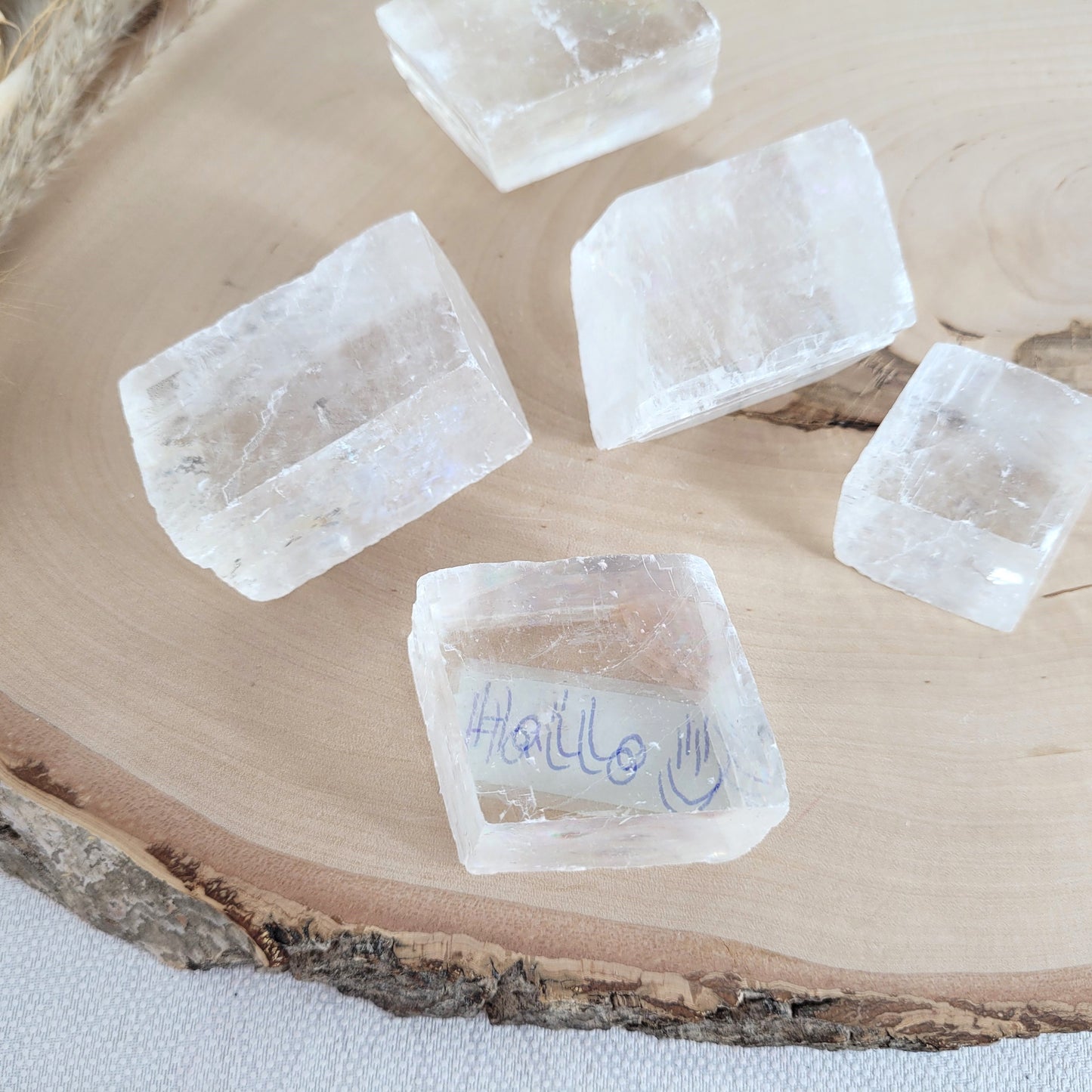 Doppelspat Kristall auf einer runden Holzscheibe präsentiert, naturbelassen mit sichtbarer Maserung – der Kristall zeigt bei Lichteinfall seine typische Doppelbrechung.