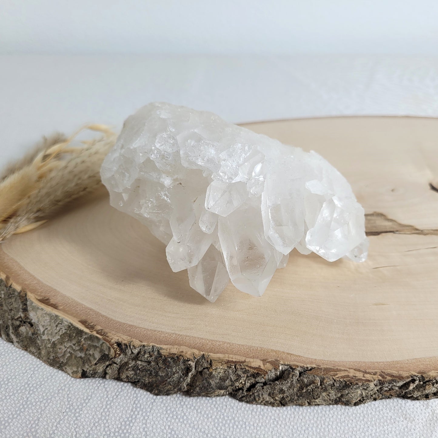 Bergkristall Naturstufe mit klaren Kristallen, platziert auf einer Holzscheibe, neutraler Hintergrund.
