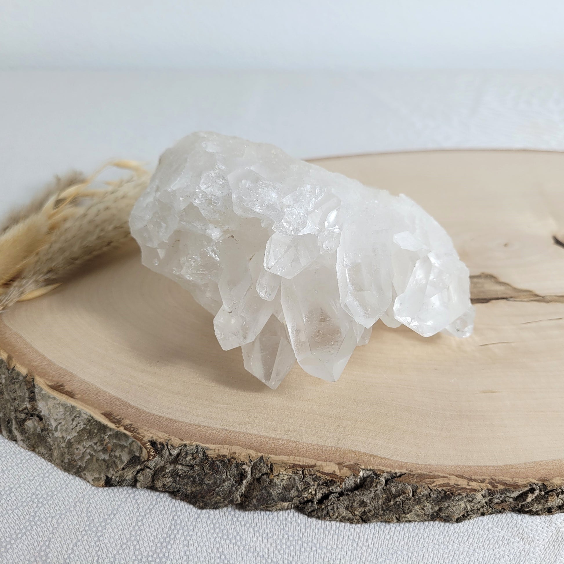 Bergkristall Naturstufe mit klaren Kristallen, platziert auf einer Holzscheibe, neutraler Hintergrund.
