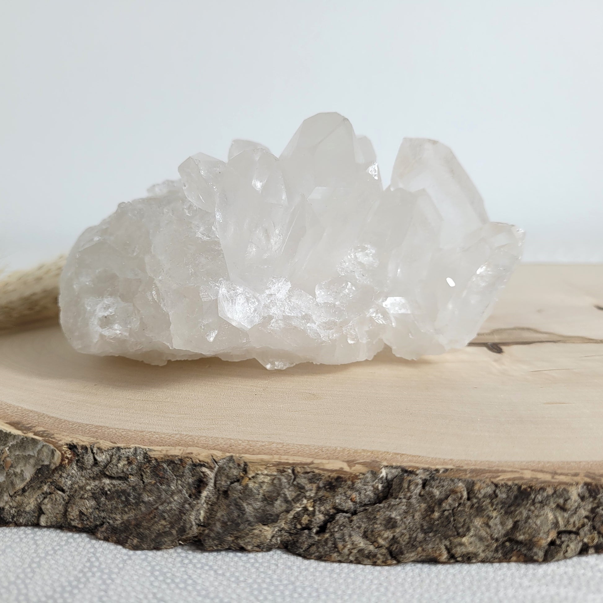Bergkristall Naturstufe mit klaren Kristallen, platziert auf einer Holzscheibe, neutraler Hintergrund.