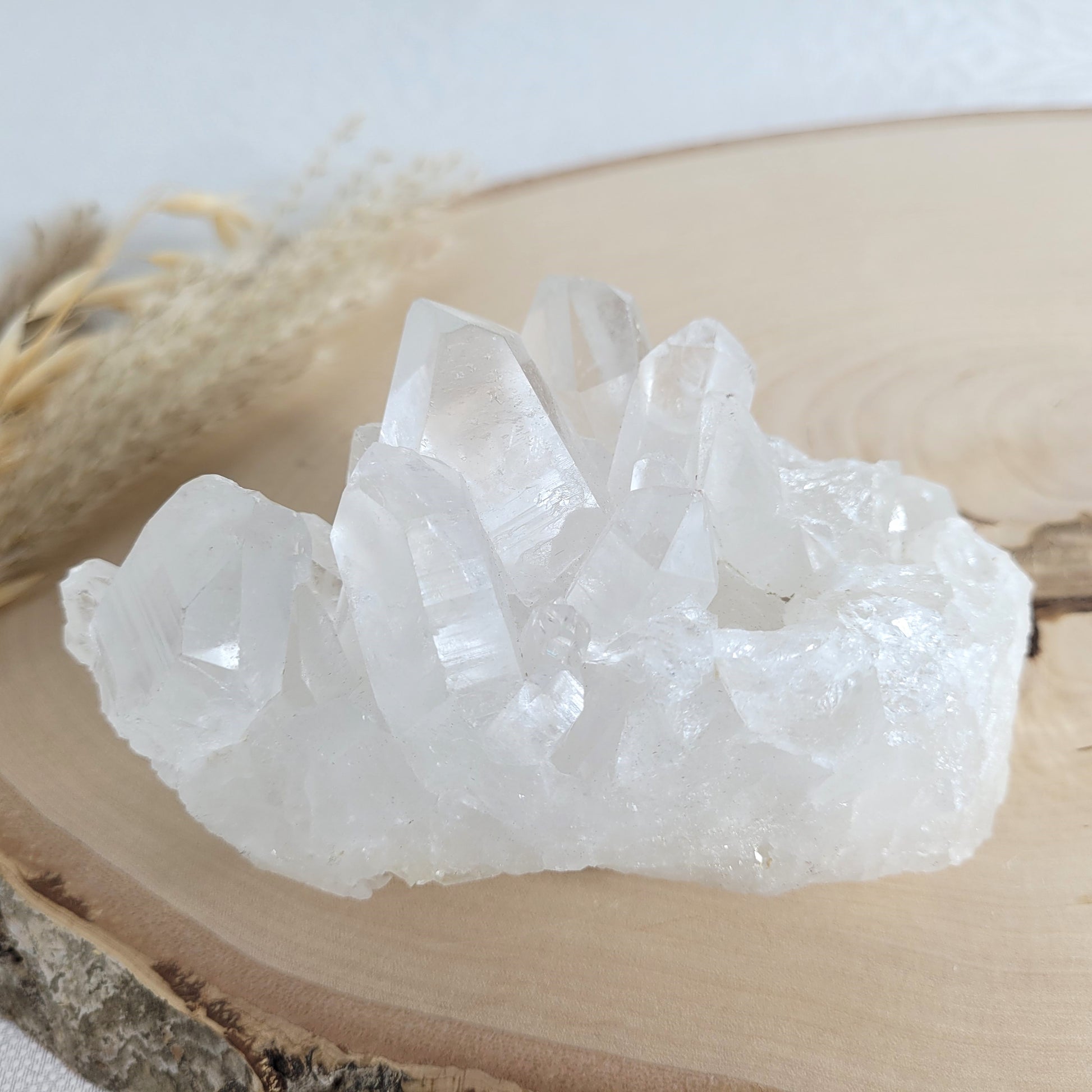 Bergkristall Naturstufe mit klaren Kristallen, platziert auf einer Holzscheibe, neutraler Hintergrund.