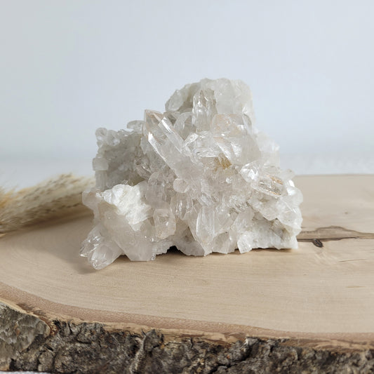 Bergkristall Naturstufe mit klaren Kristallen, platziert auf einer Holzscheibe, neutraler Hintergrund.