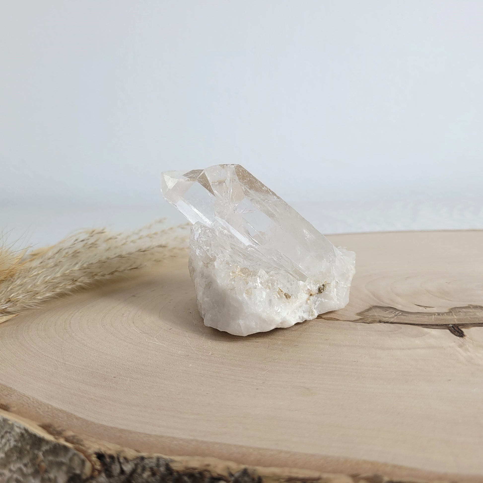 Bergkristall Naturstufe mit klaren Kristallen, platziert auf einer Holzscheibe, neutraler Hintergrund.