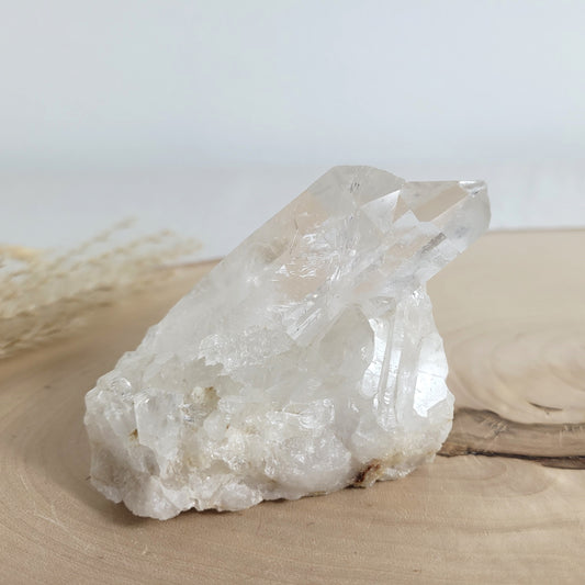 Bergkristall Naturstufe mit klaren Kristallen, platziert auf einer Holzscheibe, neutraler Hintergrund.