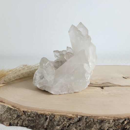 Bergkristall Naturstufe mit klaren Kristallen, platziert auf einer Holzscheibe, neutraler Hintergrund.
