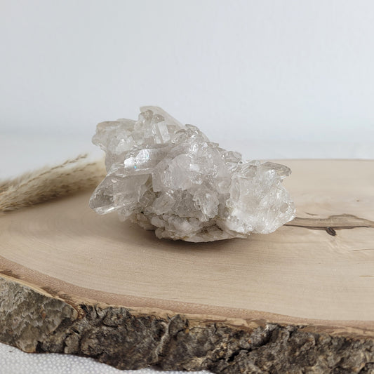 Bergkristall Naturstufe mit klaren Kristallen, platziert auf einer Holzscheibe, neutraler Hintergrund.