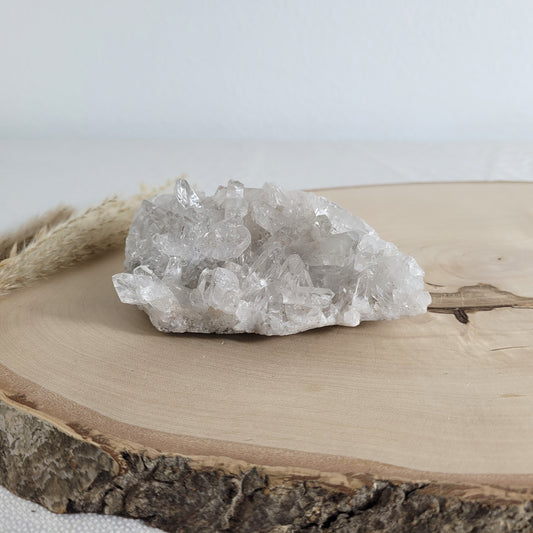 Bergkristall Naturstufe mit klaren Kristallen, platziert auf einer Holzscheibe, neutraler Hintergrund.