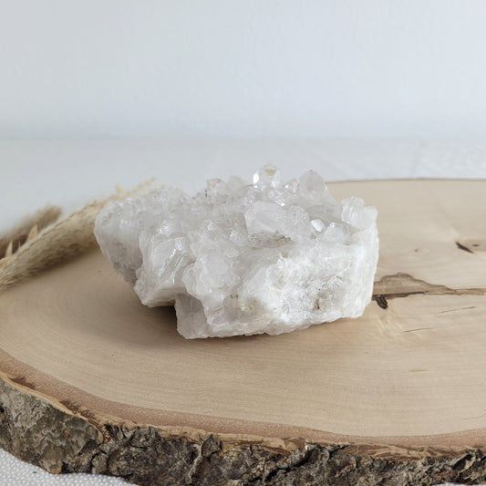 Bergkristall Naturstufe mit klaren Kristallen, platziert auf einer Holzscheibe, neutraler Hintergrund.