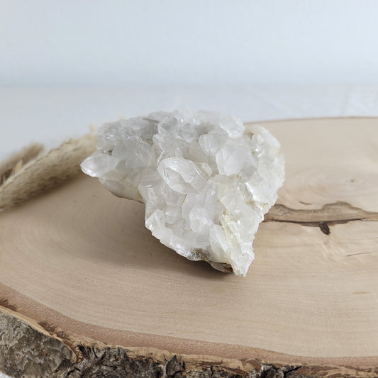 Bergkristall Naturstufe mit klaren Kristallen, platziert auf einer Holzscheibe, neutraler Hintergrund.