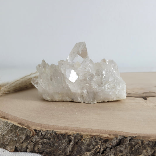 Bergkristall Naturstufe mit klaren Kristallen, platziert auf einer Holzscheibe, neutraler Hintergrund.