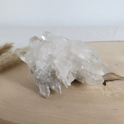 Bergkristall Naturstufe mit klaren Kristallen, platziert auf einer Holzscheibe, neutraler Hintergrund.