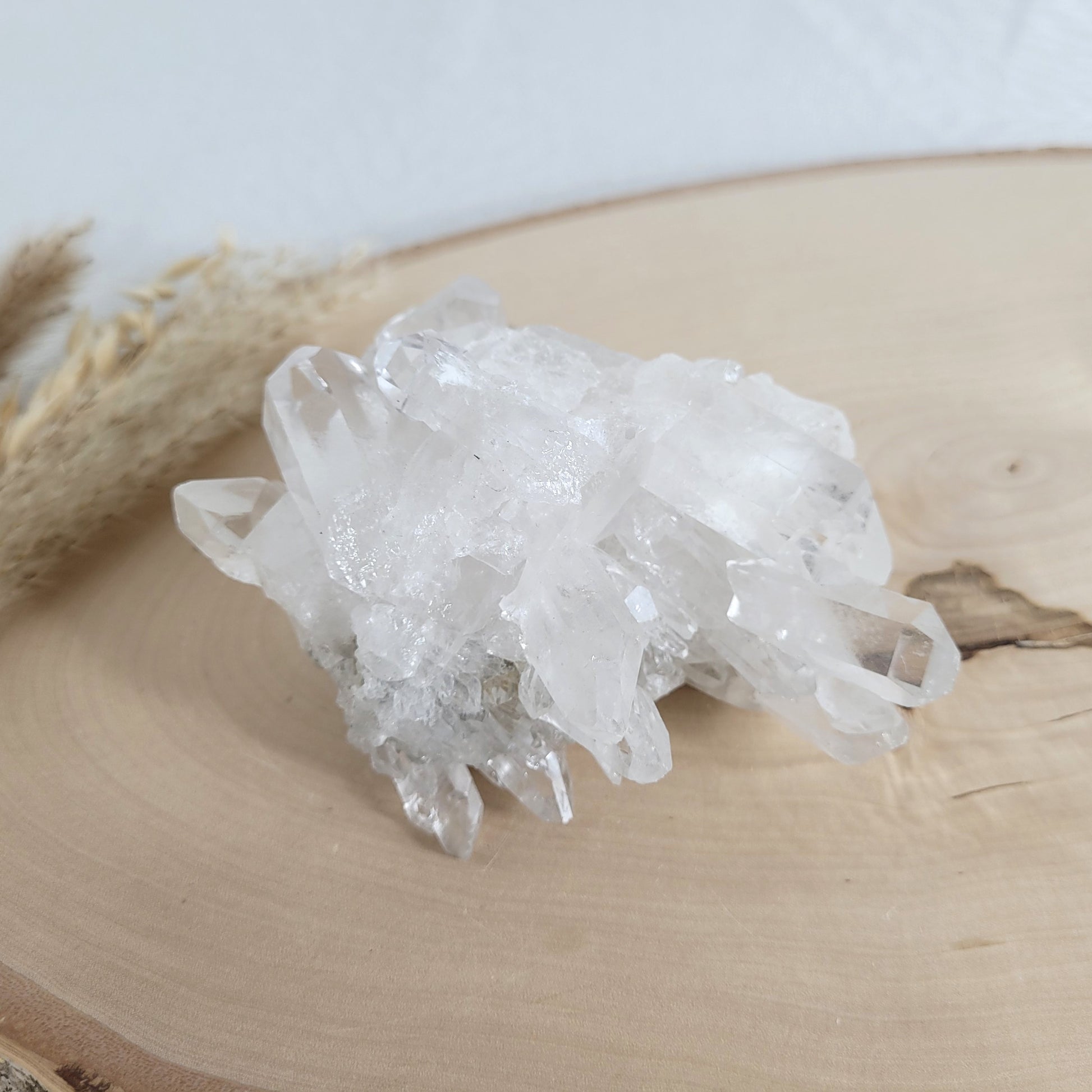Bergkristall Naturstufe mit klaren Kristallen, platziert auf einer Holzscheibe, neutraler Hintergrund.