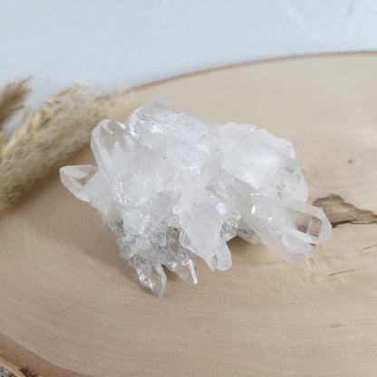 Bergkristall Naturstufe mit klaren Kristallen, platziert auf einer Holzscheibe, neutraler Hintergrund.