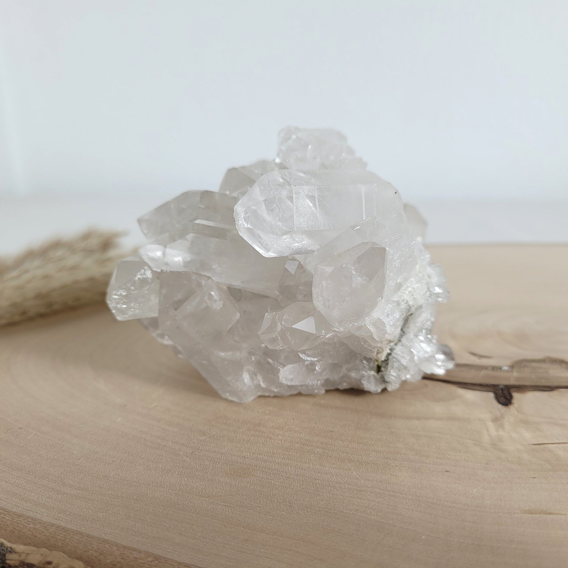 Bergkristall Naturstufe mit klaren Kristallen, platziert auf einer Holzscheibe, neutraler Hintergrund.