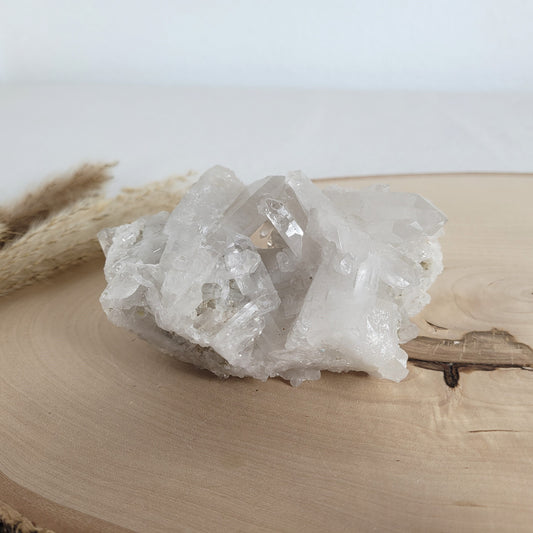 Bergkristall Naturstufe mit klaren Kristallen, platziert auf einer Holzscheibe, neutraler Hintergrund.