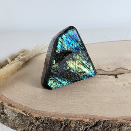 Labradorit Freiform mit natürlichem, intensivem Farbspiel, präsentiert auf einer Holzscheibe. Die typische Lamellenstruktur bricht das Licht auf besondere Weise und macht das Stück zu einem besonderen Blickfang.