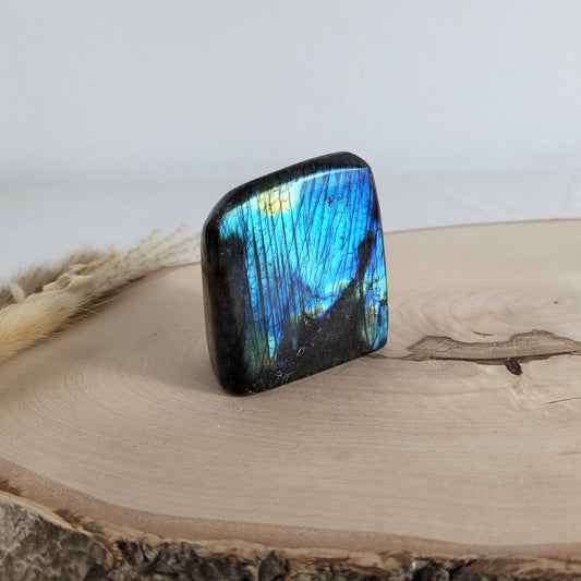 Labradorit Freiform mit natürlichem, intensivem Farbspiel, präsentiert auf einer Holzscheibe. Die typische Lamellenstruktur bricht das Licht auf besondere Weise und macht das Stück zu einem besonderen Blickfang.