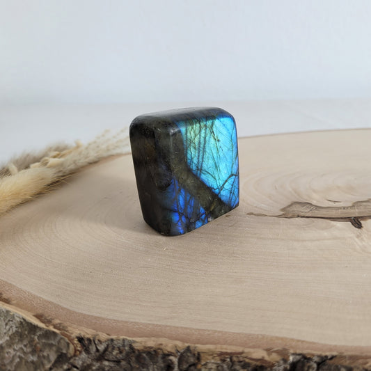 Labradorit Freiform mit natürlichem, intensivem Farbspiel, präsentiert auf einer Holzscheibe. Die typische Lamellenstruktur bricht das Licht auf besondere Weise und macht das Stück zu einem besonderen Blickfang.