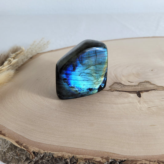 Labradorit Freiform mit natürlichem, intensivem Farbspiel, präsentiert auf einer Holzscheibe. Die typische Lamellenstruktur bricht das Licht auf besondere Weise und macht das Stück zu einem besonderen Blickfang.