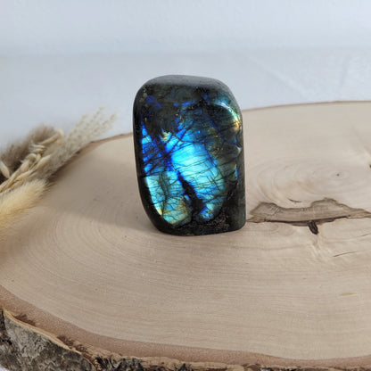 Labradorit Freiform mit natürlichem, intensivem Farbspiel, präsentiert auf einer Holzscheibe. Die typische Lamellenstruktur bricht das Licht auf besondere Weise und macht das Stück zu einem besonderen Blickfang.