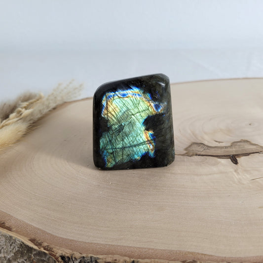 Labradorit Freiform mit natürlichem, intensivem Farbspiel, präsentiert auf einer Holzscheibe. Die typische Lamellenstruktur bricht das Licht auf besondere Weise und macht das Stück zu einem besonderen Blickfang.
