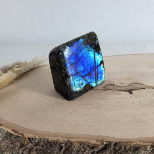 Labradorit Freiform mit natürlichem, intensivem Farbspiel, präsentiert auf einer Holzscheibe. Die typische Lamellenstruktur bricht das Licht auf besondere Weise und macht das Stück zu einem besonderen Blickfang.