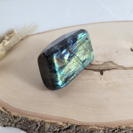 Labradorit Freiform mit natürlichem, intensivem Farbspiel, präsentiert auf einer Holzscheibe. Die typische Lamellenstruktur bricht das Licht auf besondere Weise und macht das Stück zu einem besonderen Blickfang.