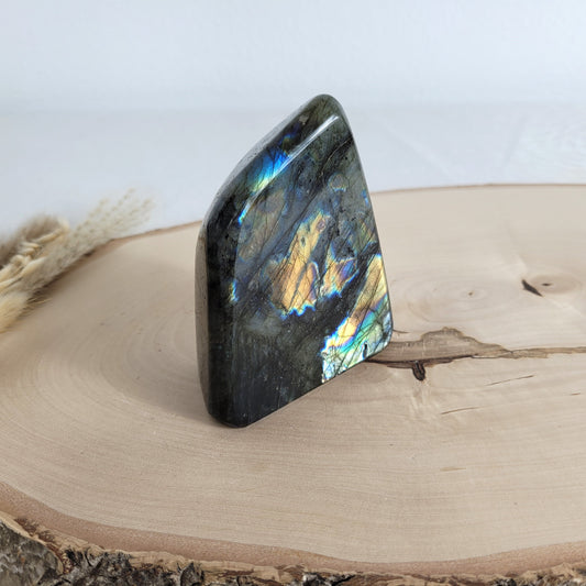 Labradorit Freiform mit natürlichem, intensivem Farbspiel, präsentiert auf einer Holzscheibe. Die typische Lamellenstruktur bricht das Licht auf besondere Weise und macht das Stück zu einem besonderen Blickfang.