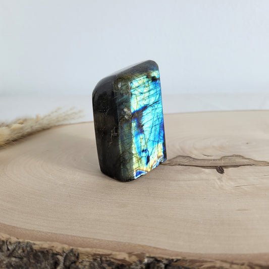 Labradorit Freiform mit natürlichem, intensivem Farbspiel, präsentiert auf einer Holzscheibe. Die typische Lamellenstruktur bricht das Licht auf besondere Weise und macht das Stück zu einem besonderen Blickfang.