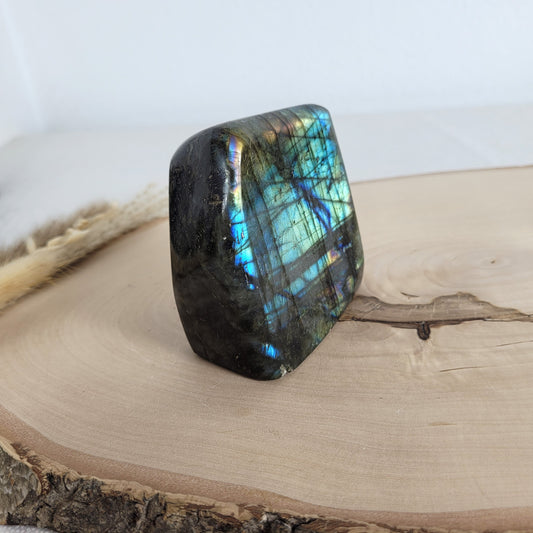 Labradorit Freiform mit natürlichem, intensivem Farbspiel, präsentiert auf einer Holzscheibe. Die typische Lamellenstruktur bricht das Licht auf besondere Weise und macht das Stück zu einem besonderen Blickfang.