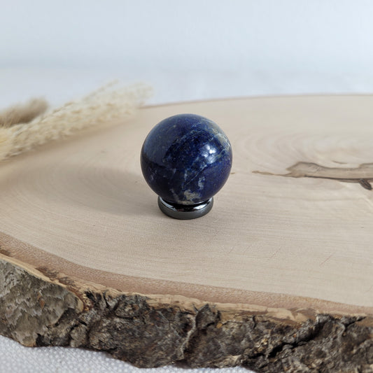 Lapis Lazuli Kugel mit intensiver blauer Farbe und goldglänzenden Pyrit-Einschlüssen, dekorativ platziert auf einer natürlichen Holzscheibe.